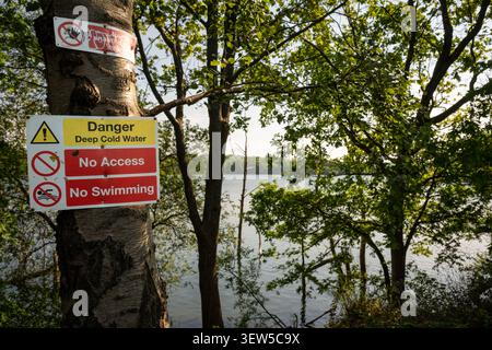 Danger sign warning of deep cold water. No access. No swimming. Warning sign on a tree with trees and lake behind. Stock Photo
