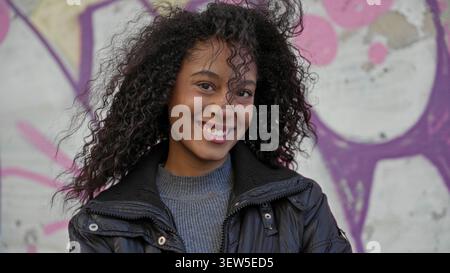 Teen woman smiling, teeth visible, wearing jacket and gray sweater against graffiti wall on street; youthful joy. Stock Photo