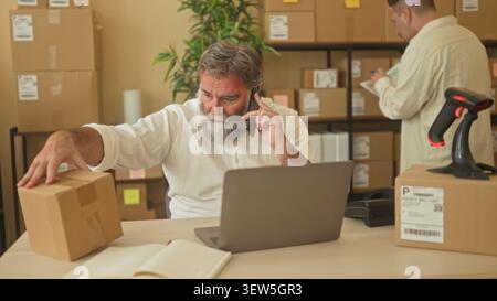 Two men with hand on parcel, holding phone to ear and leaning over laptop in building; teamwork productivity. Stock Photo