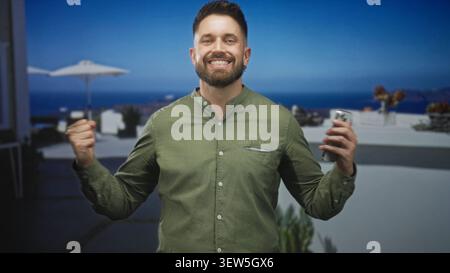 Bearded hispanic man tapping smartphone screen with thumb while smiling, holding phone in left hand on building terrace overlooking sea; joy connectio Stock Photo