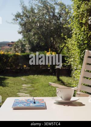 White table with coffee, notebook and pen, blurry Mediterranean garden with olive tree in background, vertical Stock Photo