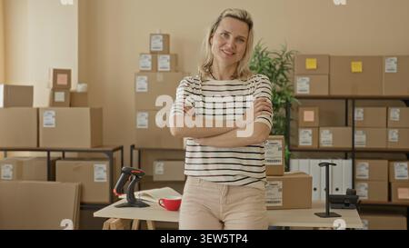 Woman with crossed arms beside packages on a packing table inside a building, barcode scanner and stacked boxes visible; confidence entrepreneurship. Stock Photo