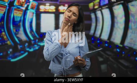 Thai woman holding clipboard touching neck in a casino building with slot machines and neon lights; fatigue contemplation. Stock Photo