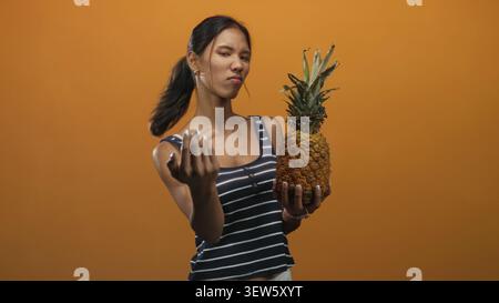 Thai woman brunette wearing navy striped tank holds a ripe pineapple in left hand and beckons with right palm forward on an orange studio set; skeptic Stock Photo