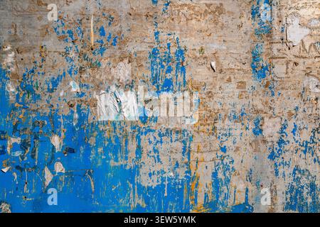 Close-up shot of a weathered wall with peeling blue paint and exposed concrete. The surface displays a grunge aesthetic with scattered paper fragments Stock Photo