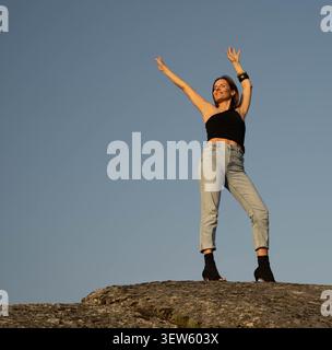 Woman standing on a rock with arms raised against a clear sky Stock Photo