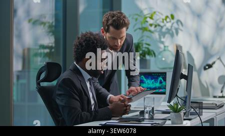Two multiethnic business men working in office. African American leader work with computer talking advise to Caucasian manager asking help with Stock Photo