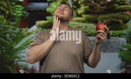 Man holding a red apple with hand on cheek for toothache in forest path wearing brown t shirt; discomfort. Stock Photo
