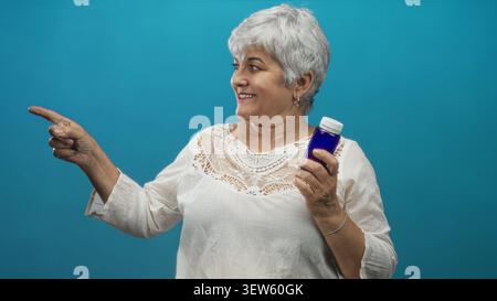 Senior woman with grey hair and plus size holding a blue medicine bottle points finger and smiles in studio; wellness confidence. Stock Photo