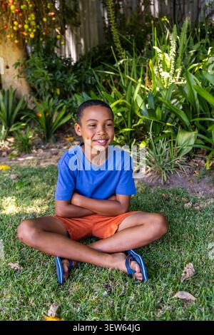 African American child sitting cross-legged on lawn wearing blue T-shirt, orange shorts, flip-flops Stock Photo