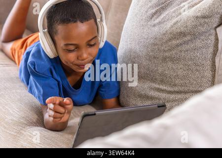 African American child lying on couch in blue tee and orange shorts wearing headset tapping tablet Stock Photo