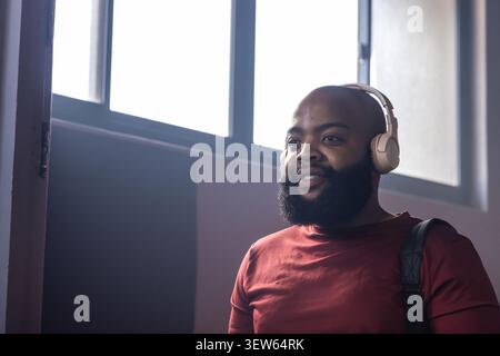 Man standing in corridor by windows wearing maroon tee, headphones and backpack, copy space Stock Photo