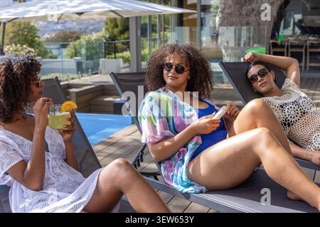 Diverse female friends reclining on pool loungers in cover-ups, sipping cocktail and holding phone Stock Photo
