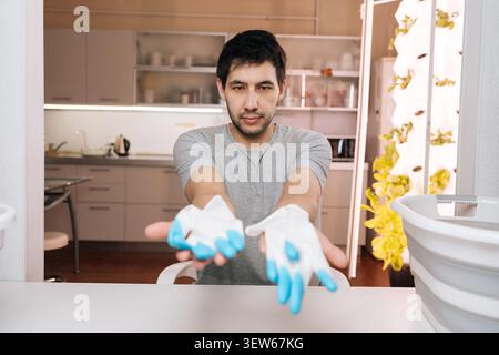 Young man offers gardening gloves to camera. He sitting at table with modern kitchen and vertical hydroponic farm in background. Concept of urban Stock Photo