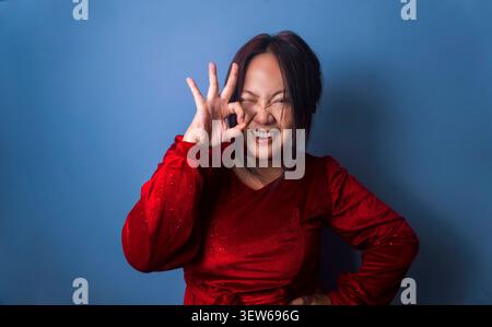 Smiling young woman in a red velvet dress making an OK hand gesture over one eye while posing against a blue studio background. Stock Photo