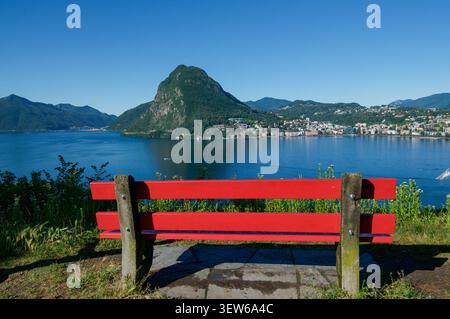 Scenic wide panorama of Lake Lugano and Monte San Salvatore peak, featuring an empty red park bench in the foreground. Captured from the viewpoint of Stock Photo
