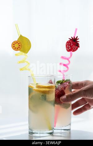 Hand placing two tall glasses of refreshing lemonade and strawberry drinks with fruit-themed straws, illustrating delicious, cool, and healthy beverag Stock Photo