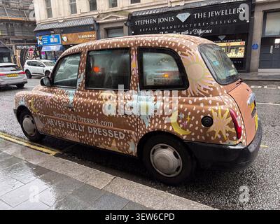 A London-style taxi wrapped in Never Fully Dressed branding parked on a wet street in Liverpool, UK. Stock Photo