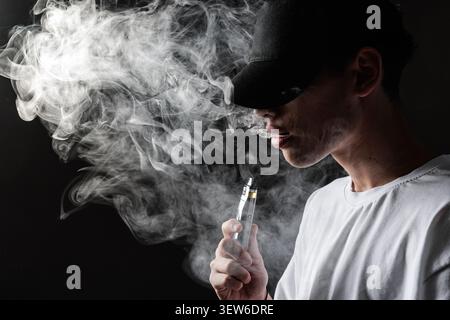 A young man with a cap exhales vapor from an e-cigarette, highlighting the dangers of vaping and nicotine addiction in a moody atmosphere. Stock Photo