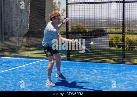 Male wearing grey shirt and black shorts swinging paddle, hitting yellow ball on blue turf court Stock Photo