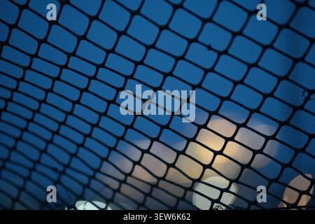 Macro view of a plastic mesh fence creating a geometric pattern against a blue evening sky with soft out-of-focus city lights Stock Photo