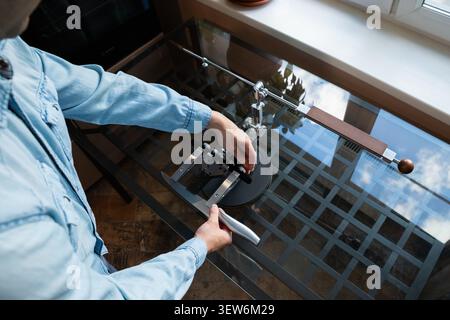 Man fastens a kitchen knife into a precision sharpener. Male user setting up blade in a sharpening tool on a glass table. Equipment for professional t Stock Photo