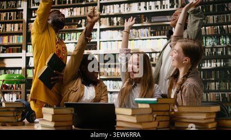 Smiling diverse students stacking hands together in unity at library table, celebrating teamwork and successful group study session. Classmates united in academic work. Stock Photo