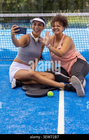 African American female duo taking smartphone selfie sitting on blue court with paddles and ball Stock Photo