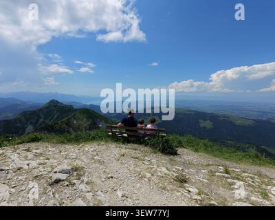 Aerial view of a man and two children sitting on a wooden bench on a mountain peak overlooking the vast alpine landscape in Gruyeres, Fribourg, Switze Stock Photo