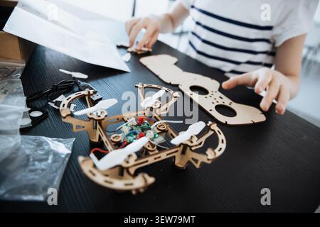 A smart girl works on assembling a DIY wooden drone in her living room. She uses a remote control and follows steps for STEM learning and engineering. Stock Photo
