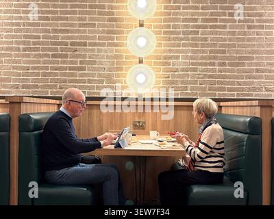 Elderly couple sitting in a booth using a tablet and smartphone at a dining table with brick wall background. Stock Photo