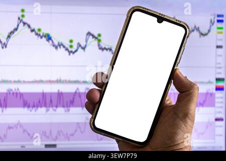 A person's hand holds a mobile phone with an isolated white screen for mockup space, positioned in front of a computer monitor displaying stock market Stock Photo