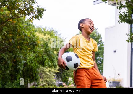 Child in yellow tee, orange shorts, laughing, holding black-white soccer-ball in yard, copy space Stock Photo
