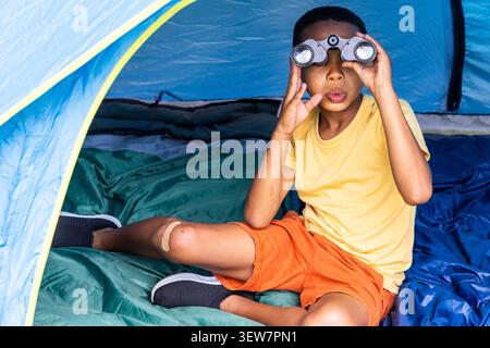 African American boy sitting inside blue tent holding black binoculars in yellow tee orange shorts Stock Photo