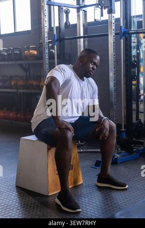 African American man sitting on wooden plyometric box leaning forward in gym near kettlebells Stock Photo