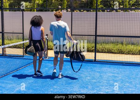 Couple in sportswear walking on blue racket court toward mesh fence carrying duffels and rackets Stock Photo