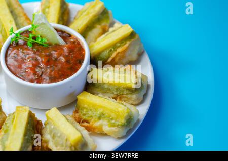 Golden fried green peppers served with spicy sauce. Bright blue background enhances vibrant food colors. Slices look crisp and savory, perfect for sha Stock Photo