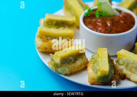 Golden fried green peppers served with spicy sauce. Bright blue background enhances vibrant food colors. Slices look crisp and savory, perfect for sha Stock Photo