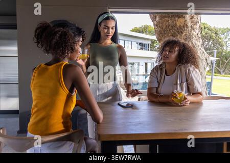 Three diverse female friends sitting at patio wooden table holding juice, one reaching smartphone Stock Photo