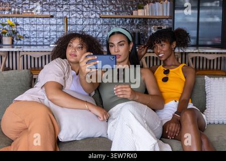 Three women sitting on green sofa at cafe taking selfie with blue phone while wearing sunglasses Stock Photo