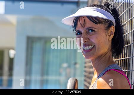 Mature woman of African descent leaning on metal fence, holding paddle, wearing visor and grey tank Stock Photo