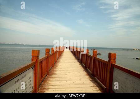 A wooden boardwalk stretches out over the calm, blue sea, with light-colored planks and rich reddish-brown railings creating a warm, inviting contrast Stock Photo