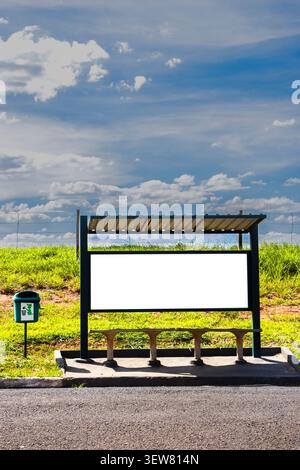 A blank white billboard stands inside a rustic public bus shelter on a sunny day in rural Brazil. The scene features a green trash can, simple concret Stock Photo