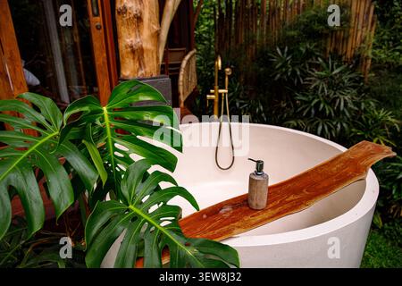 aesthetic outdoor white round bathtub with gold faucet in bali jungle with big green leaves Stock Photo