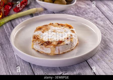 Baked Camembert cheese on white plate with golden brown rind melted center. Background features green olives red chili peppers leek on rustic gray woo Stock Photo