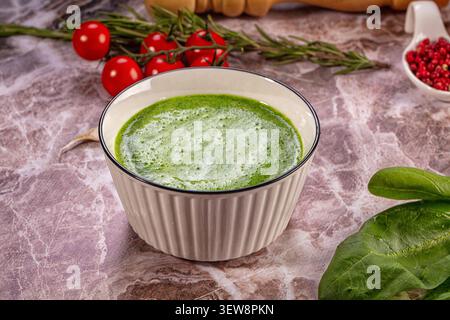 Vibrant green soup in ribbed ceramic bowl surrounded by fresh spinach leaves cherry tomatoes rosemary sprigs pink peppercorns on textured marble Stock Photo