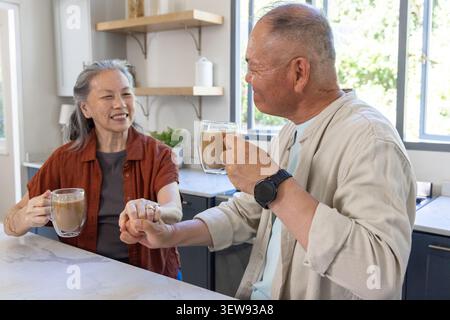 Senior Asian couple sitting at kitchen counter holding mugs clasping hands, wearing rust beige tops Stock Photo