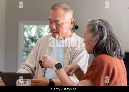 Senior Asian couple sitting at dining table using tablet and holding card while checking smartwatch Stock Photo