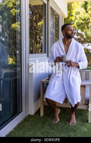 Mature African man sitting on balcony bench holding ceramic mug by sliding door, wearing bathrobe Stock Photo