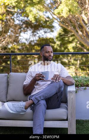 Mature African American man sitting on cushioned patio sofa holding ceramic mug, wearing T-shirt Stock Photo